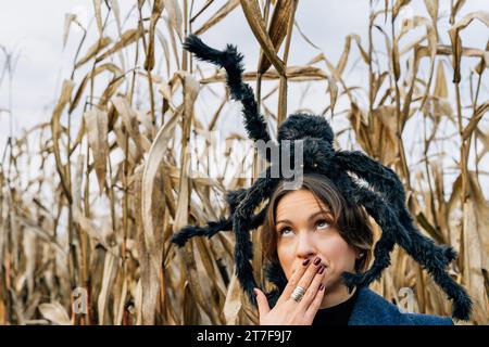 Une femme couvre sa bouche avec sa main dans la peur à cause d'une grosse araignée jouet sur sa tête. Costumes et décorations d'Halloween Banque D'Images