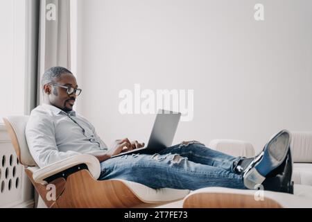 Homme d'affaires détendu dans des lunettes à l'aide d'un ordinateur portable, confortable dans une chaise moderne Banque D'Images
