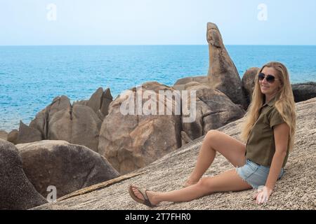 Une femme aux cheveux longs est assise sur une pierre au bord de la mer avec pour toile de fond le monument Grandparents Rock sur Koh Samui en Thaïlande. Banque D'Images