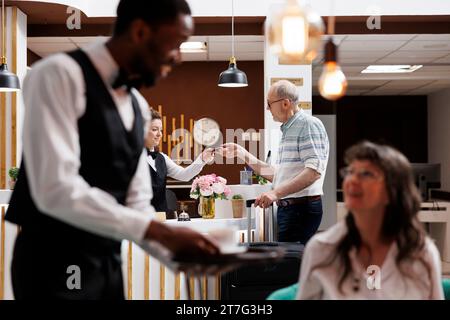 Le réceptionniste caucasien aide le vieil homme retraité avec l'enregistrement à l'hôtel vérifiant l'identité et récupérant les détails de réservation. Le concierge féminin tient et vérifie le passeport du touriste masculin. Concentrez-vous sur l'arrière-plan. Banque D'Images