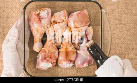Les mains du chef portant des gants versent de la sauce soja dans un plat de cuisson en verre avec des pilons de poulet dedans. Recette de viande de poulet marinée Banque D'Images