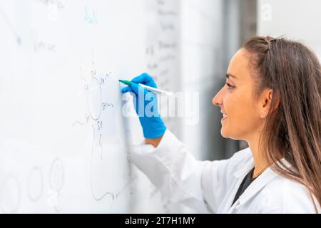 Vue latérale d'une jeune femme biologiste utilisant un tableau blanc pour écrire des données dans un laboratoire Banque D'Images