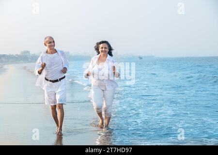 Heureux couple indien senior portant des vêtements blancs courir ensemble à la plage et s'amuser. Vacances et concept de santé Banque D'Images