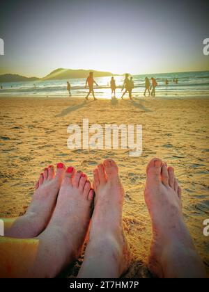 Un couple se repose sur Patong Beach à Phuket, Thaïlande au coucher du soleil avec seulement leurs pieds visibles, les gens et la mer en arrière-plan floues. Banque D'Images