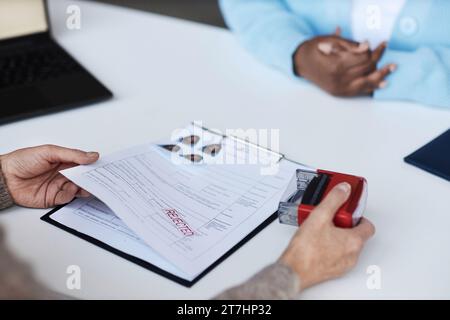 Mains de la gestionnaire féminine du centre de demande de visa mettant le timbre rejeté sur le formulaire rempli du demandeur afro-américain assis devant elle Banque D'Images