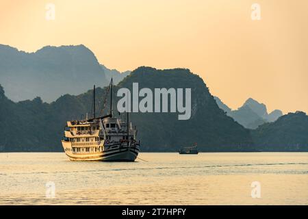 Un bateau de pêche traditionnel vietnamien squd passe derrière un grand bateau de croisière touristique au coucher du soleil sur la baie d'Halong, Vietnam Banque D'Images