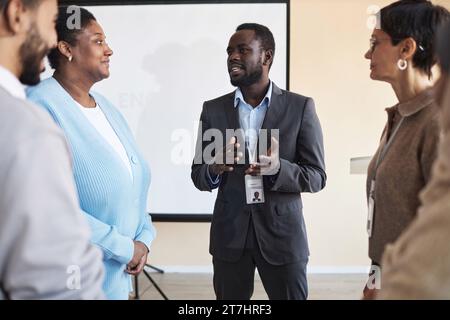 Jeune homme noir confiant en costume parlant à un public interculturel tout en se tenant devant le tableau blanc après une formation commerciale dans la salle de conférence Banque D'Images