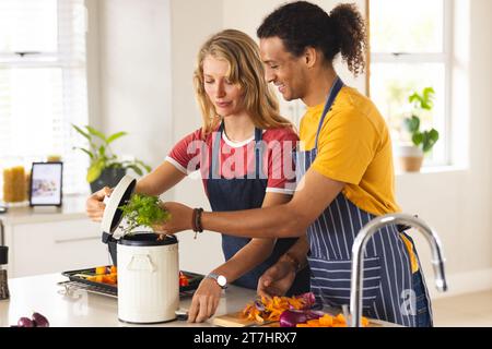 Heureux couple diversifié compostage des déchets végétaux dans la cuisine ensoleillée à la maison Banque D'Images