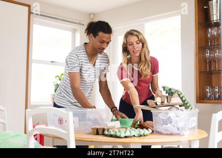 Heureux couple diversifié triant les déchets pour le recyclage dans la cuisine ensoleillée à la maison Banque D'Images