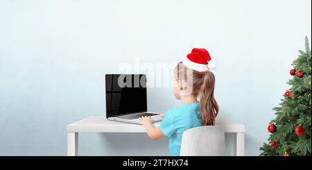 Fille avec chapeau de Noël rouge travaille sur ordinateur portable dans la chambre avec arbre de vacances. Soncept d'écrire des cartes de voeux et des lettres au Père Noël Banque D'Images