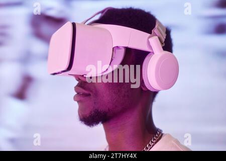 Portrait vue de côté d'un jeune homme afro-américain portant un casque VR dans une lumière néon rose entrant dans le cyberespace Banque D'Images