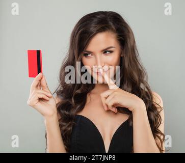 Glamour joyeuse modèle brunette femme tenant la carte de crédit regardant la caméra isolée sur fond gris de bannière murale studio Banque D'Images