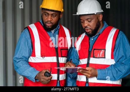 Portrait de deux ingénieurs ou contremaîtres africains portent un EPI vérifiant le stockage du conteneur avec le fond du conteneur de fret au coucher du soleil. Logistique importation globale ou Banque D'Images