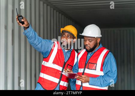 Portrait de deux ingénieurs ou contremaîtres africains portent un EPI vérifiant le stockage du conteneur avec le fond du conteneur de fret au coucher du soleil. Logistique importation globale ou Banque D'Images