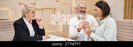 agent immobilier d'âge moyen regardant un couple marié heureux tenant la clé de la nouvelle maison dans le bureau, bannière Banque D'Images