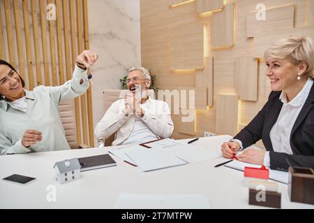 femme heureuse tenant la clé de la nouvelle maison près excité mari d'âge moyen et agent immobilier dans le bureau Banque D'Images