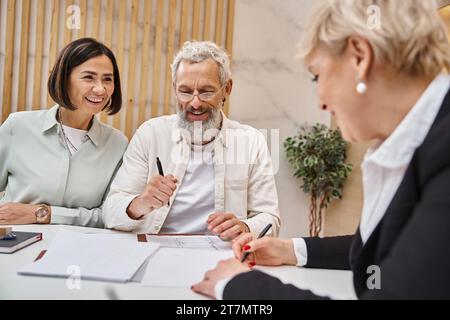 heureux agent immobilier d'âge moyen signant contact près du couple marié dans le bureau immobilier, vente maison Banque D'Images