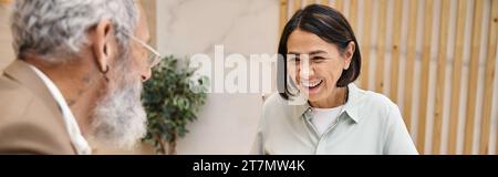 heureuse femme d'âge moyen souriant près de l'agent immobilier barbu dans le bureau immobilier, bannière Banque D'Images