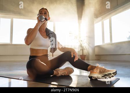 Femme assise sur tapis de yoga dans la salle de gym, eau potable, salle ensoleillée, relaxation Banque D'Images