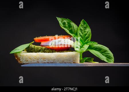 tranche de pain fraîche avec sauce pesto, mozzarella et tomate sur une spatule en bois avec basilic. Isolé sur fond noir. Banque D'Images