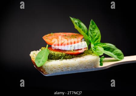 tranche de pain fraîche avec sauce pesto, mozzarella et tomate sur une spatule en bois avec basilic. Isolé sur fond noir. Banque D'Images