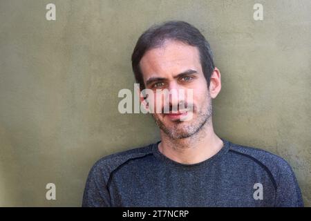 Portrait en plein air d'un jeune homme caucasien de 30 ans avec une barbe courte, des cheveux noirs et des yeux bleus souriant à la caméra contre un mur vert plat. Copier spa Banque D'Images