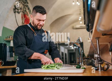 Chef cuisinier barbu vêtu d'uniforme noir avec un tablier chopiant concombre vert frais avec couteau tranchant dans la cuisine du restaurant du sous-sol. Professionnel Banque D'Images