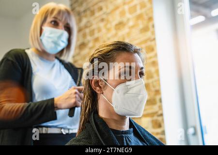 Coiffeur dans un masque peignant les cheveux mouillés d'un client dans un salon pendant COVID-19 Banque D'Images