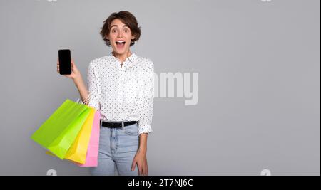 Jeune femme souriante avec des sacs à provisions montre son téléphone intelligent sur la caméra. Black Friday, consommateur, acheteur, vente, magasin, paiement, achat en ligne, remise, Banque D'Images