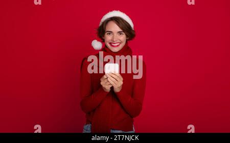 Heureuse femme mignonne dans le chapeau de Père Noël rouge utilise smartphone pour surfer sur Internet ou discuter avec bien-aimé ou ami. Joyeux noël. Bonne année. Holida Banque D'Images