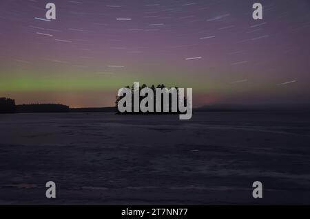 Sentiers étoilés et teinte verte des aurores boréales lointaines au-dessus d'une île boisée dans un lac gelé en hiver en Finlande Banque D'Images