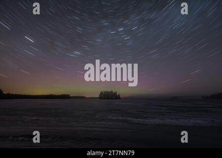 Sentiers étoilés et teinte verte des aurores boréales lointaines au-dessus d'une île boisée dans un lac gelé en hiver en Finlande Banque D'Images