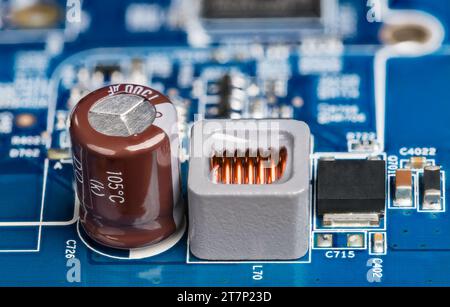 Gros plan de l'inductance, du condensateur électrolytique ou du transistor sur fond de carte flou. Carte de circuit imprimé d'alimentation bleue avec composants électroniques. Banque D'Images