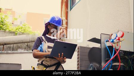 Technicien certifié travaillant sur le système cvc extérieur, tenant un ordinateur portable. Mécanicien compétent optimisant les performances du climatiseur externe, assurant qu'il fonctionne à sa capacité maximale Banque D'Images