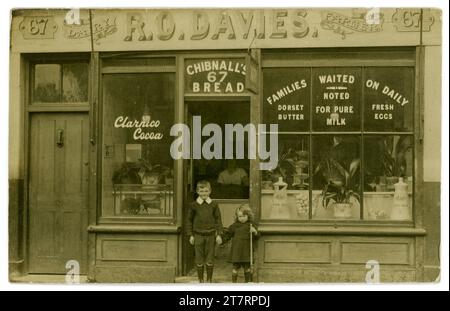 Carte postale originale d'un garçon mignon souriant et de son frère (qui tient un fusil jouet) debout à l'extérieur de la boutique de ses parents au-dessus de lui est un signe pour R O Davies Dairy Farmer. Le magasin vend principalement des produits laitiers et la vitrine du magasin fait de la publicité pour le beurre Dorset, le lait pur et les œufs et que «les familles sont attendues quotidiennement». Circa 1915 de la coiffure de Lady Shopkeeper. Il y a une publicité pour le pain de Chibnall au-dessus de la porte. La boulangerie de Chibnall était une entreprise londonienne. ROYAUME-UNI Banque D'Images