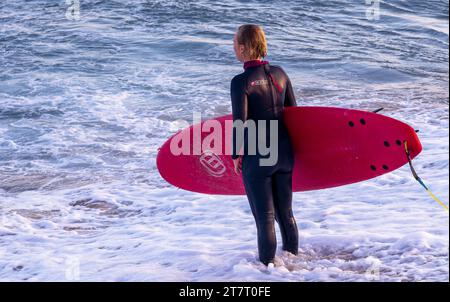 Femme mature en tenue de surf avec un tableau rouge contemple le coucher du soleil sur la plage de Barcelone, en Espagne. Banque D'Images