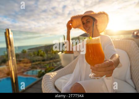 Femme dans un chapeau de soleil profitant d'un cocktail à l'hôtel tropical de plage sur une terrasse ensoleillée avec une piscine en arrière-plan Banque D'Images