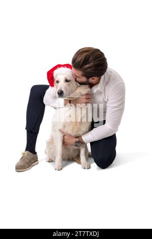 Beau jeune homme avec chien blanc labrador portant le chapeau rouge du Père Noël pour célébrer la saison, sur fond blanc en studio Banque D'Images