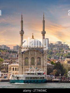 Istanbul, Turquie - Mai 8 2023 : vue depuis le détroit du Bosphore surplombant la mosquée Ortakoy, ou Ortakoy Camii, alias Buyuk Mecidiye Camii avec un ferry devant le coucher du soleil, adapté à la place de la jetée d'Ortakoy Banque D'Images