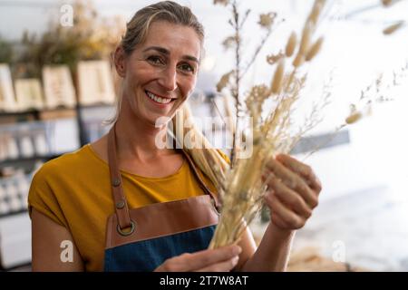 Une fleuriste souriante tient un bouquet de blé séché dans un atelier lumineux Banque D'Images