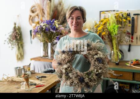 Femme avec des lunettes souriant et tenant une grande couronne florale séchée dans un atelier d'artisanat Banque D'Images