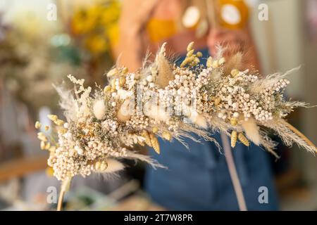Gros plan d'une couronne de fleurs séchées tenue par une personne dans un tablier bleu, fond flou Banque D'Images
