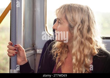 Daydreasing Commute : une jeune femme de 30 ans, aux cheveux blonds et vernis à ongles rouge, regarde sans hésiter par la fenêtre du train pendant son voyage quotidien, Banque D'Images