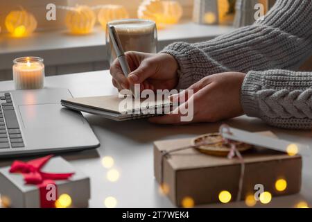 Jeune femme écrivant des cartes de noël, fait l'ordre sur ordinateur portable. Femme cueillant des cadeaux achats en ligne à la maison confortable parmi les boîtes-cadeaux et les paquets. Hiver s Banque D'Images