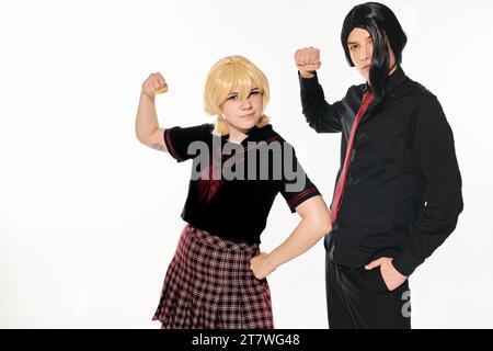 étudiants expressifs en uniforme sombre et perruques montrant les muscles et regardant la caméra sur blanc Banque D'Images