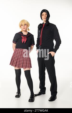 homme en vêtements noirs avec les mains dans les poches près de femme blonde en uniforme scolaire sur blanc, cosplayers Banque D'Images