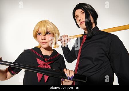 jeunes cosplayers en perruques blondes et noires posant en uniforme d'école sombre avec des battes de baseball sur gris Banque D'Images