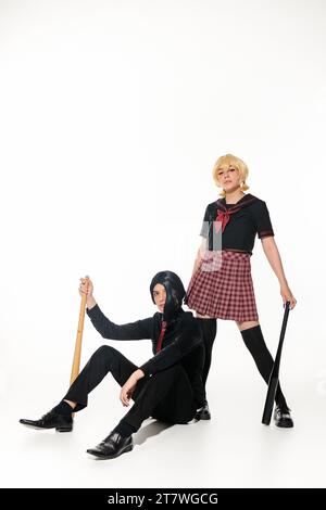 couple de style animé confiant en uniforme scolaire et perruques avec pose avec des battes de baseball sur blanc Banque D'Images