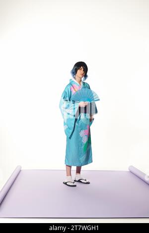 homme de style anime en kimono bleu et perruque tenant un ventilateur de main et regardant la caméra sur fond blanc Banque D'Images