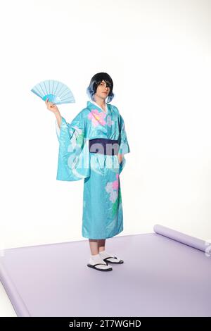 style cosplay, homme en kimono bleu et perruque posant avec ventilateur à main sur tapis violet et toile de fond blanche Banque D'Images